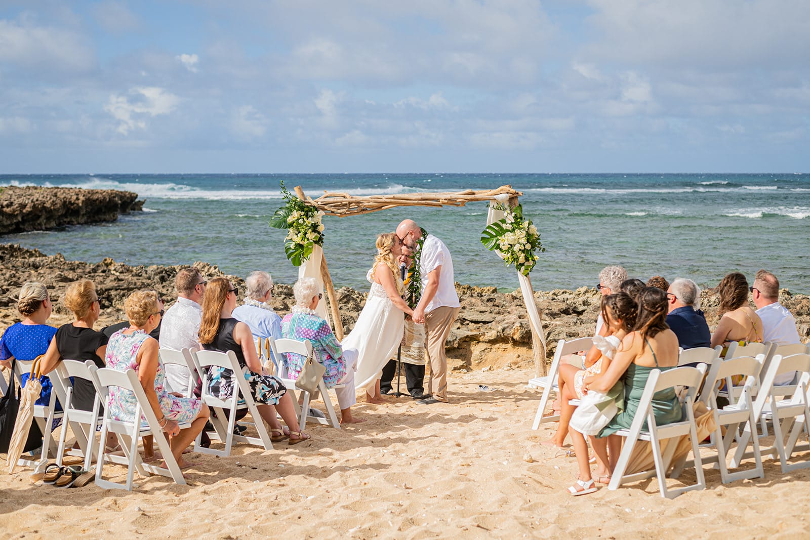 A couple's first kiss under a driftwood arch at their North Shore beach elopement ceremony on Oahu, family and friends seated on both sides