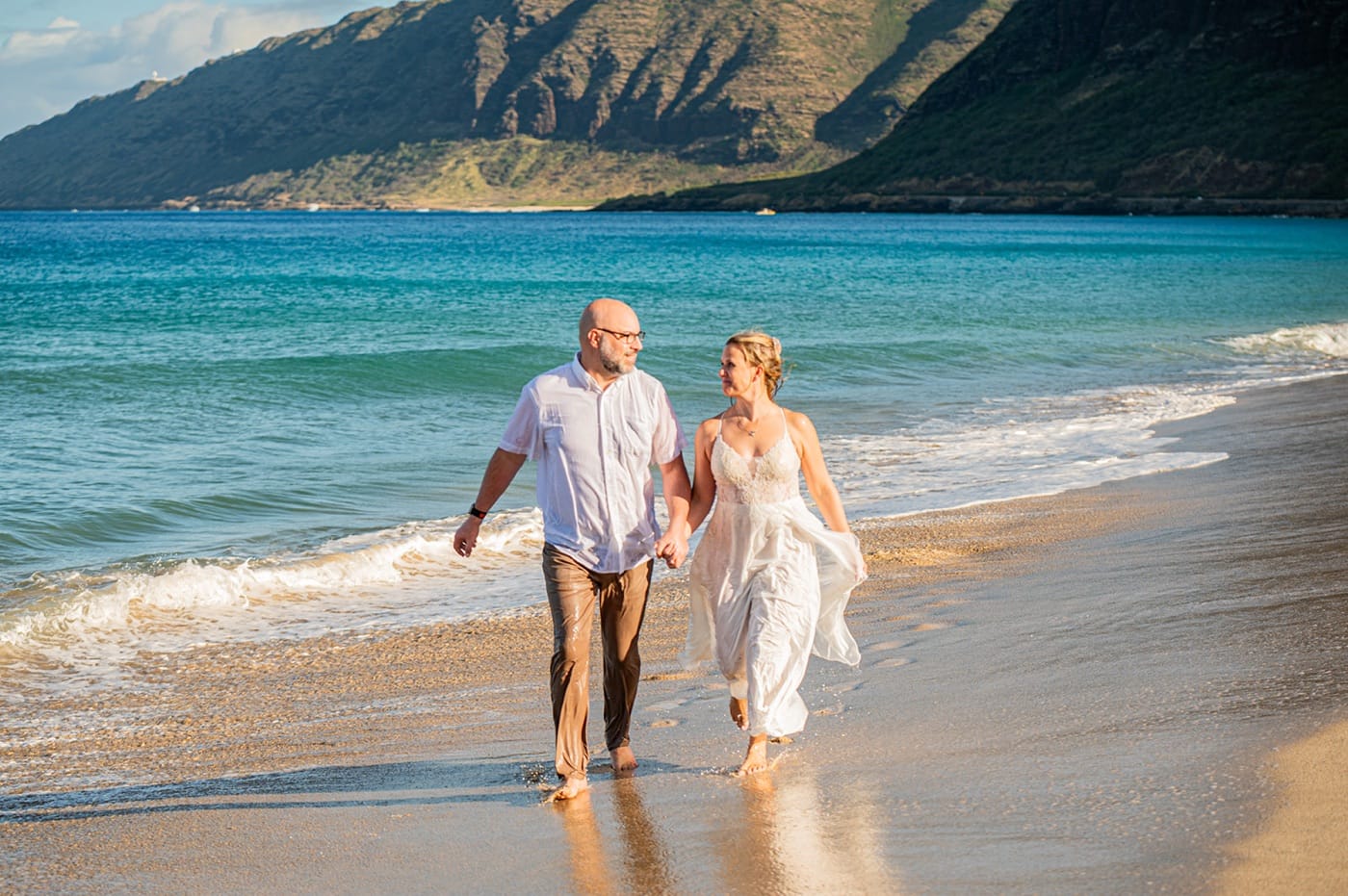 A couple walking hand in hand along the water's edge on an Oahu beach, the mountains behind them
